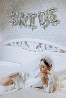 an incredibly beautiful bride in the morning in a white robe and a long veil lies on the bed in her bedroom near gel balls with the inscription bride. The bride poses in the morning. photo