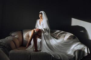 an incredibly beautiful bride in the morning in a white robe and a long veil sits on the bed in her bedroom. The bride poses in the morning before the wedding ceremony. photo