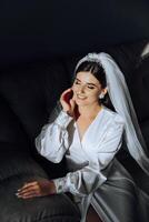 retrato de un increíblemente hermosa niña novia en un blanco túnica en el dormitorio, el novia poses en el Mañana antes de el Boda ceremonia. foto