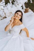 The brunette bride, smiling sincerely, sits on the ground in a unfolded dress, near a white arch made of flowers. Wedding ceremony. Sunny day. Beautiful hair and makeup photo