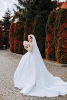 A brunette bride in a white dress with a long train holds the dress and walks down the stone path. Autumn. Wedding photo session in nature. Beautiful hair and makeup. celebration