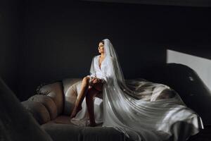 an incredibly beautiful bride in the morning in a white robe and a long veil sits on the bed in her bedroom. The bride poses in the morning before the wedding ceremony. photo