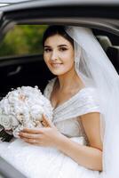 Wedding portrait. A brunette bride in an elegant dress and veil poses with a bouquet in the car. Preparation for the celebration. Beautiful hair and makeup. photo