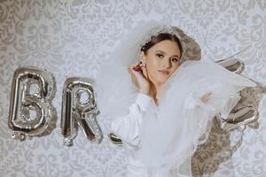 an incredibly beautiful bride in the morning in a white robe and a long veil stands on the bed in her bedroom near gel balls with the inscription bride. The bride poses in the morning. photo