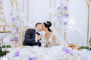 Young groom in a white shirt and smiling brunette bride at their table in a banquet hall, raising a toast and kissing against a background of fresh flowers. photo