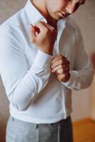 un hombre en un blanco camisa soportes por el ventana en el habitación y sujeta el botones en su collar y mangas reloj en mano. elegante negocio retrato de un hombre, de cerca foto. el novio es preparando. foto