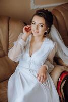 an incredibly beautiful bride in the morning in a white robe and a long veil sits on the bed in her bedroom. The bride poses in the morning before the wedding ceremony. photo