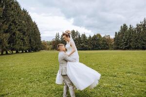 Stylish groom holds beautiful bride in long white dress in his arms and dances on green grass in autumn park, forest under sunlight. Wedding photography, portrait of smiling newlyweds. photo