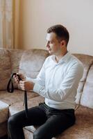 a young man in a white shirt stands by the window in the room and puts on a tie. The groom gets dressed in the morning and prepares for the wedding. photo