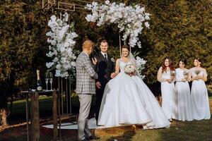 A master of ceremonies gives a speech while a bride and groom hold hands near a white floral arch photo