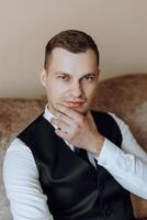 People, lifestyle, business, style, fashion and menswear concept. Positive successful young man sitting in a chair in a room, smiling at the camera, wearing elegant shoes, trousers, and a white shirt photo