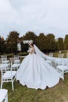 A brunette bride in a white dress with a long train, holds a bouquet and walks to the wedding arch photo