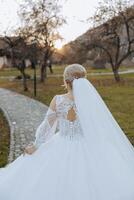 A blonde bride in a white dress with a long train holds the dress and walks along the stone path. Autumn. Wedding photo session in nature. Beautiful hair and makeup. celebration