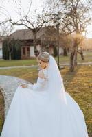 A blonde bride in a white dress with a long train holds the dress and walks along the stone path. Autumn. Wedding photo session in nature. Beautiful hair and makeup. celebration