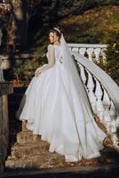 A brunette bride in a white dress with a long train holds the dress and walks down the stone path. Autumn. Wedding photo session in nature. Beautiful hair and makeup. celebration