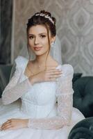 fashion photo of a beautiful bride with dark hair in an elegant wedding dress and stunning makeup in the room on the morning of the wedding. The bride is preparing for the wedding