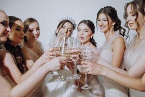 Photo with bridesmaids drinking champagne from glasses in the morning in a beautiful hotel. Photo of a beautiful young bride and her friends in matching dresses drinking champagne before the wedding.