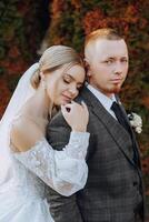 encantador y elegante recién casados son abrazando y sonriente en contra el antecedentes de otoño naturaleza en un hermosa jardín. un increíblemente hermosa joven novia inclinado en contra el hombro de su amado novio foto