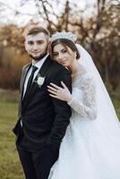 Stylish, young groom and beautiful bride in a long white dress and a long veil with a bouquet in their hands, hugging in the park in the autumn nature. Wedding portrait of newlyweds. photo