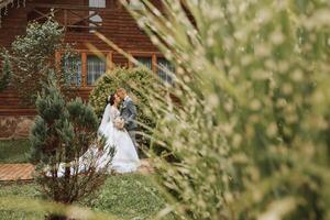 Handsome groom and charming bride stay together near modern wooden house in park. Wide-angle photo, place for text photo