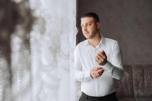 un hombre en un blanco camisa soportes por el ventana en el habitación y sujeta el botones en su collar y mangas reloj en mano. elegante negocio retrato de un hombre, de cerca foto. el novio es preparando. foto