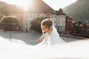 Rizado rubia novia en un blanco vestido, cubierto con un velo, poses para el cámara con un ramo de flores de rosas. retrato de el novia. hermosa maquillaje y cabello. Boda en naturaleza foto