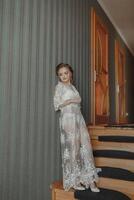 A delicate wedding dress hangs on a beautiful railing in the hallway of the house. The bride is standing on the steps in a delicate transparent petticoat. Nice light. Wedding photo. A high quality photo