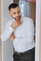 un hombre en un blanco camisa soportes por el ventana en el habitación y sujeta el botones en su collar y mangas reloj en mano. elegante negocio retrato de un hombre, de cerca foto. el novio es preparando. foto