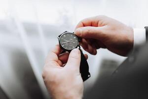 A close-up of a cropped frame of a man puts on a watch with a leather belt, is dressed in a stylish suit, a white shirt, wears a gold ring. photo