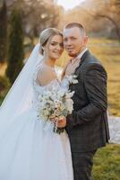 Stylish, young groom and beautiful bride in a long white dress and a long veil with a bouquet in their hands, hugging in the park in the autumn nature. Wedding portrait of newlyweds. photo