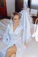 an incredibly beautiful bride in the morning in a white robe and a long veil sits on the bed in her bedroom. The bride poses in the morning before the wedding ceremony. photo