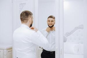 A man in a white shirt stands by the window in the room and fastens the buttons on his collar and sleeves. Watch on hand. Stylish business portrait of a man, close-up photo. The groom is preparing. photo