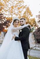 elegante, joven novio y hermosa novia en un largo blanco vestir y un largo velo con un ramo de flores en su manos, abrazando en el parque en el otoño naturaleza. Boda retrato de recién casados. foto