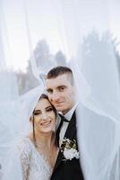 Stylish, young groom and beautiful bride in a long white dress and a long veil with a bouquet in their hands, hugging in the park in the autumn nature. Wedding portrait of newlyweds. photo