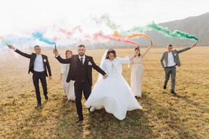 Wedding photo session in nature. Bride and groom and their friends in a field, cheerfully holding colored smoke.