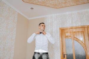 un joven hombre en un blanco camisa soportes por el ventana en el habitación y pone en un atar. el novio obtiene vestido en el Mañana y prepara para el boda. foto