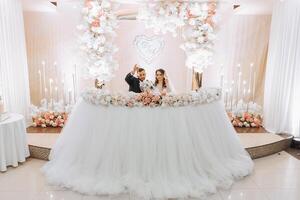 un joven novio en un negro traje y un sonriente morena novia son a su mesa en un banquete sala, levantamiento un brindis en contra un antecedentes de Fresco flores foto