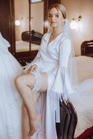 an incredibly beautiful bride in the morning in a white robe and a long veil sits on the bed in her bedroom. The bride poses in the morning before the wedding ceremony. photo