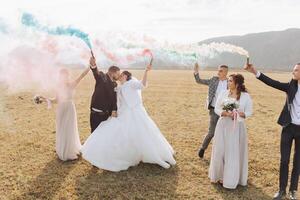 Wedding photo session in nature. Bride and groom and their friends in a field, cheerfully holding colored smoke.