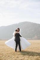 elegante novio sostiene hermosa novia en largo blanco vestir en su brazos y bailes en verde césped en otoño parque, bosque debajo luz de sol. Boda fotografía, retrato de sonriente recién casados. foto