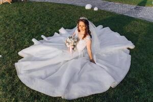 A brunette bride with a tiara in her hair, sitting on the ground in a dress unfolded in the shape of a circle, holding a bouquet. On a green background. Sunny day. Wedding ceremony photo
