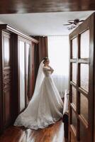 Young brunette in long wedding dress and veil in hotel room. A charming bride, full length, in a magnificent white dress on the morning before the wedding. photo