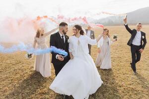 Wedding photo session in nature. Bride and groom and their friends in a field, cheerfully holding colored smoke.