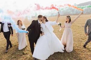 Wedding photo session in nature. Bride and groom and their friends in a field, cheerfully holding colored smoke.