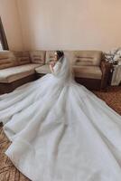 An incredibly beautiful bride sits on a bed in her bedroom. Bride morning. A gorgeous bride in a beautiful dress is sitting on a bed in a bright room . photo