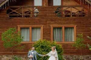 Wedding in nature. Photo of the bride and groom on a walk against the background of trees, the bride and groom holding hands and looking at each other against the background of trees and a wooden hut.