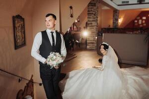 Wedding portrait of the bride and groom. Indoor photography with beautiful light. A stylish groom in a black vest is standing in front of the bride, holding a bouquet, the bride is sitting behind. photo