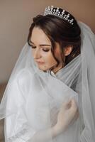 retrato de un increíblemente hermosa niña novia en un blanco túnica en el dormitorio, el novia poses participación su velo en su manos y cubre sí misma con él. foto