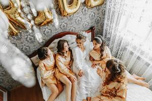 Young bridesmaids in silk robes in a hotel room. Beautiful women celebrating a bachelorette party sitting on the bed in themed pajamas. photo