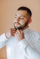 un hombre en un blanco camisa soportes por el ventana en el habitación y sujeta el botones en su collar y mangas reloj en mano. elegante negocio retrato de un hombre, de cerca foto. el novio es preparando. foto